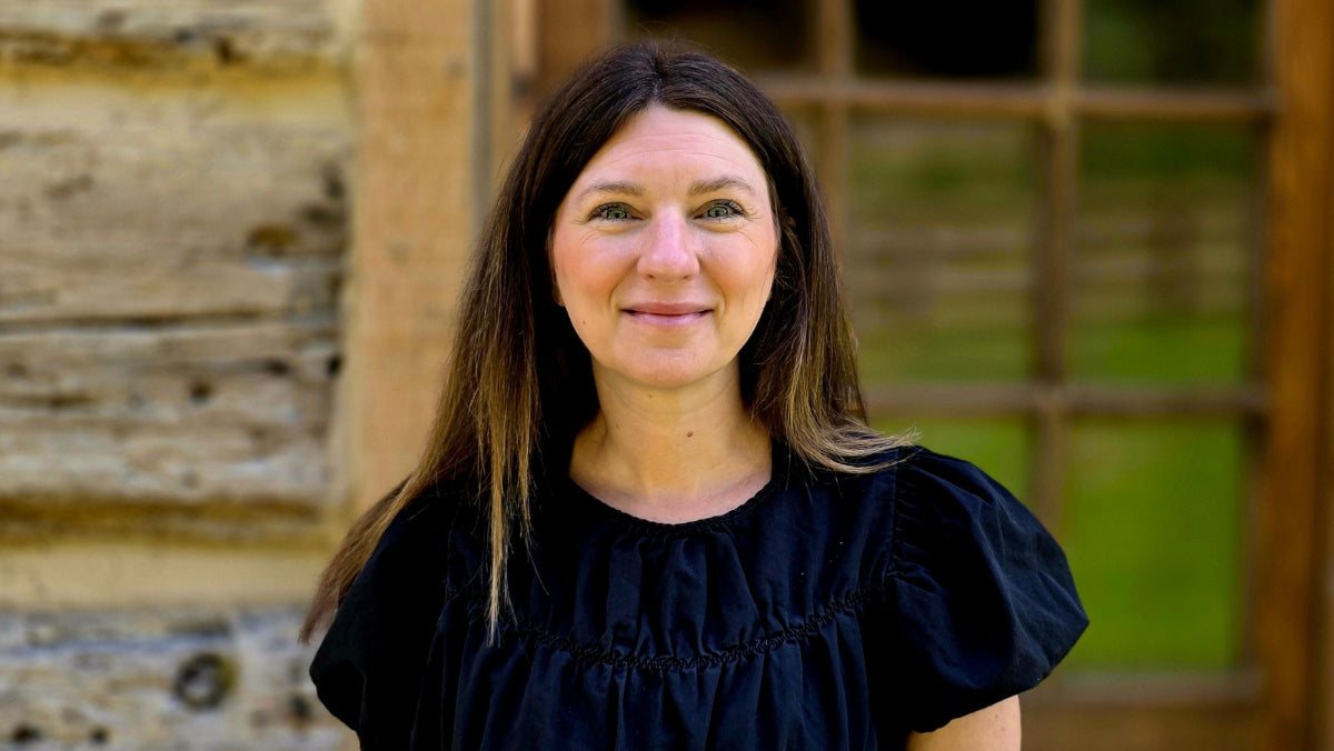 A woman with long brown hair wearing a black short-sleeved blouse stands in front of a rustic wooden cabin door.