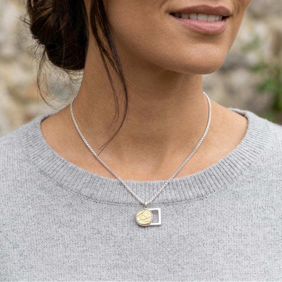 A close-up lifestyle shot of a woman wearing a delicate silver chain necklace with a pendant. The pendant features a gold-toned circular coin charm on the left and a polished silver geometric frame on the right.